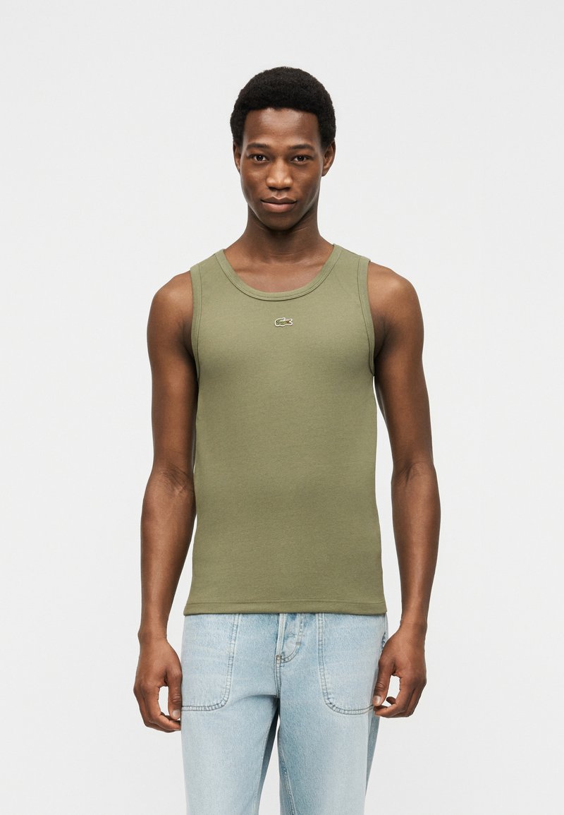 Νεαρός άνδρας που φοράει αμάνικο τοπ ελιάς με μικρό λογότυπο και ανοιχτόχρωμα τζιν, στέκεται μπροστά σε απλό ανοιχτό φόντο.