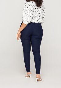 Navyblauwe, nauwsluitende broek met een gladde textuur en een hoge taille, gecombineerd met een witte blouse met stippen. Gekenmerkt door open teen witte hoge hakken.