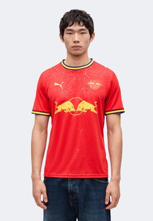 Hombre con camiseta roja de fútbol del RB Leipzig con logos amarillos de toros, emblema de Puma y patrón de hojas, combinado con vaqueros azules, de pie frente a un fondo liso.