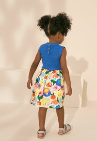 Haut en tricot bleu avec des manches courtes et une fermeture à trois boutons, associé à une jupe à motif floral coloré et des sandales bleu clair.