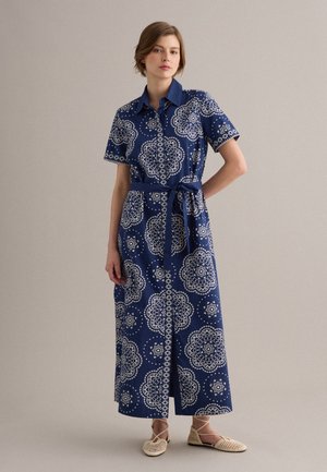 Donna che indossa un abito camicia maxi blu con motivi geometrici bianchi, maniche corte, cintura e scarpe basse beige intrecciate.