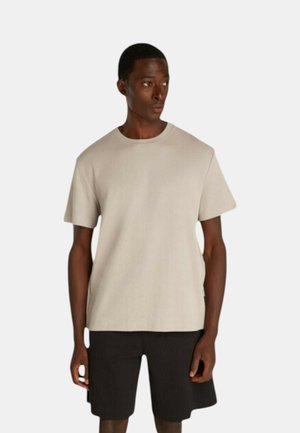 T-shirt beige a maniche corte con colletto rotondo, texture a costine, forma boxy, abbinato a pantaloni neri. Design minimalista, senza motivi visibili o accessori.