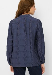 Blusa blu navy con motivo a onde testurizzato, maniche lunghe e scollatura arricciata. Realizzata in tessuto leggero con una finitura semi-trasparente.