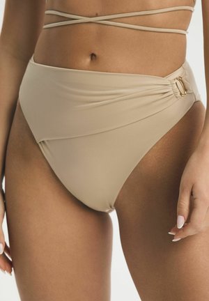 Béžové bikiny s vysokým pásom, detailom na zavinutie a zlatým krúžkom, nosené s rovnako sfarbenými prekríženými ramienkami vrchnej časti bikin na brušku.
