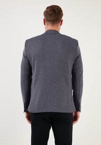 Mężczyzna stoi plecami do kamery, ubrany w szary żakiet i czarne spodnie, na tle jasnego, jednolitego tła.