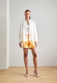Camicia bianca a maniche lunghe con una vestibilità rilassata sopra un top senza maniche bianco a costine. Pantaloni corti gialli con motivi floreali e accenti arancioni, abbinati a tacchi beige.