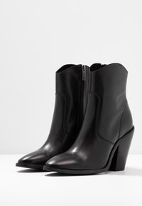 Botas de piel negras hasta el tobillo con punta afilada y un tacón de bloque de 10 cm. Presentan un cierre de cremallera lateral y una textura suave sin patrones visibles.