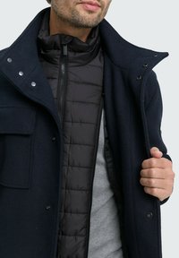 Palton din lână de culoare marină, cu un guler înalt, detalii cu capse și o vestă neagră din puf cu fermoar în interior, peste o cămașă gri. Material texturat.