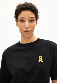 Zwarte katoenen t-shirt met een ronde halslijn, voorzien van een kleine gele strik geborduurd op de linker borstzijde.