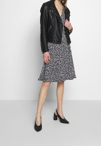 Czarna skórzana kurtka motocyklowa noszona na białej i czarnej sukience w wzór, uzupełniona czarnymi szpilkami. Sukienka ma rozkloszowany dół.