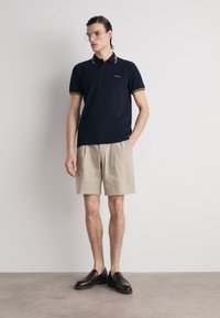 Polo shirt bleu marine avec des accents jaunes, shorts beiges et mocassins foncés. Le mannequin se tient avec les mains dans les poches, portant des lunettes transparentes dans un cadre neutre.