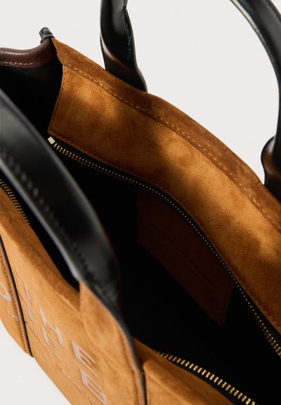 Barna velúr kézitáska fekete bőr fogantyúkkal, nyitott tetejű cipzárral és látható belső zsebbel, amelynek díszítő varrása van.