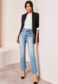 Blazer negro ajustado sobre una camiseta blanca, combinado con jeans de talle alto de color azul claro y tacones beige con acentos negros. Texturas suaves.