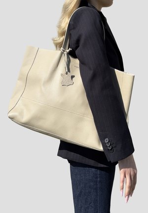 Donna con capelli biondi lunghi che indossa un blazer nero a righe sottili e jeans scuri, mentre porta una grande borsa tote in pelle beige sulla spalla.
