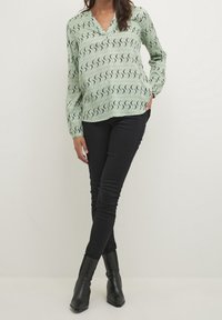 Vaaleanvihreä pusero mustalla aaltoilevalla kuviolla, V-kaulus ja pitkät hihat; yhdistetty mustiin kapealestisiin farkkuihin ja mustiin nilkkureihin.