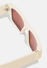 Occhiali da sole con una montatura in plastica color crema, forma conica e lenti rosse sfumate. Dettagli in oro sull'arto. Finitura opaca.