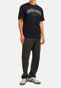 Marineblaues T-Shirt mit "JACK & JONES" Grafik in Weiß, kombiniert mit dunkelgrauen Jeans und beigen Turnschuhen mit schwarzen Akzenten.