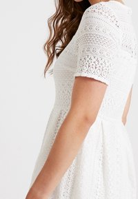 Biała sukienka koronkowa z krótkimi rękawami, skomplikowanymi wzorami, dopasowanym topem i rozkloszowanym dołem. Teksturowany i lekki materiał.