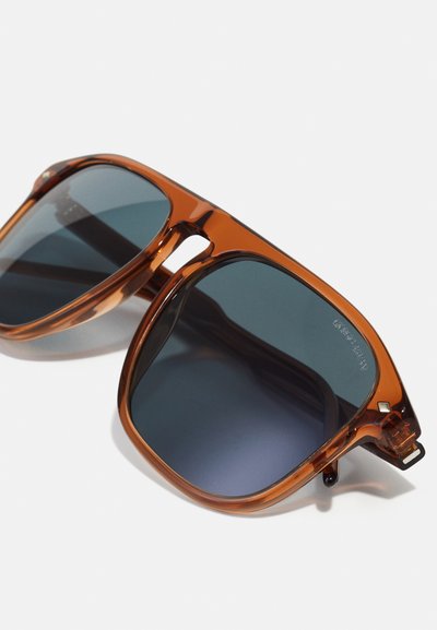 Lunettes de soleil avec une monture en plastique transparent marron et des verres teintés gris. Forme carrée avec des branches épaisses et des accents matériels minimes.