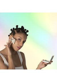 Modèle aux cheveux texturés en petits chignons tenant un applicateur de beauté vert et blanc contre son visage. Elle tient également un petit miroir compact.