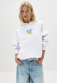 Donna bionda che indossa una felpa leggera grigia con grafica e testo di mango, abbinata a jeans larghi blu chiaro e una cintura borchiata, le mani nelle tasche.