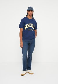 Morska majica s žutim natpisom "BERKELEY" i grafikom; plave traperice; bijele tenisice s smešnim đonom; traper kapa. Jednostavan, ležeran outfit.