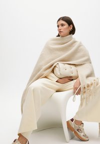Beige uld poncho med frynsekant, båret over et kridhvidt outfit, mens der holdes en hvid håndtaske med guld accenter, kombineret med beige og maroon sneakers.