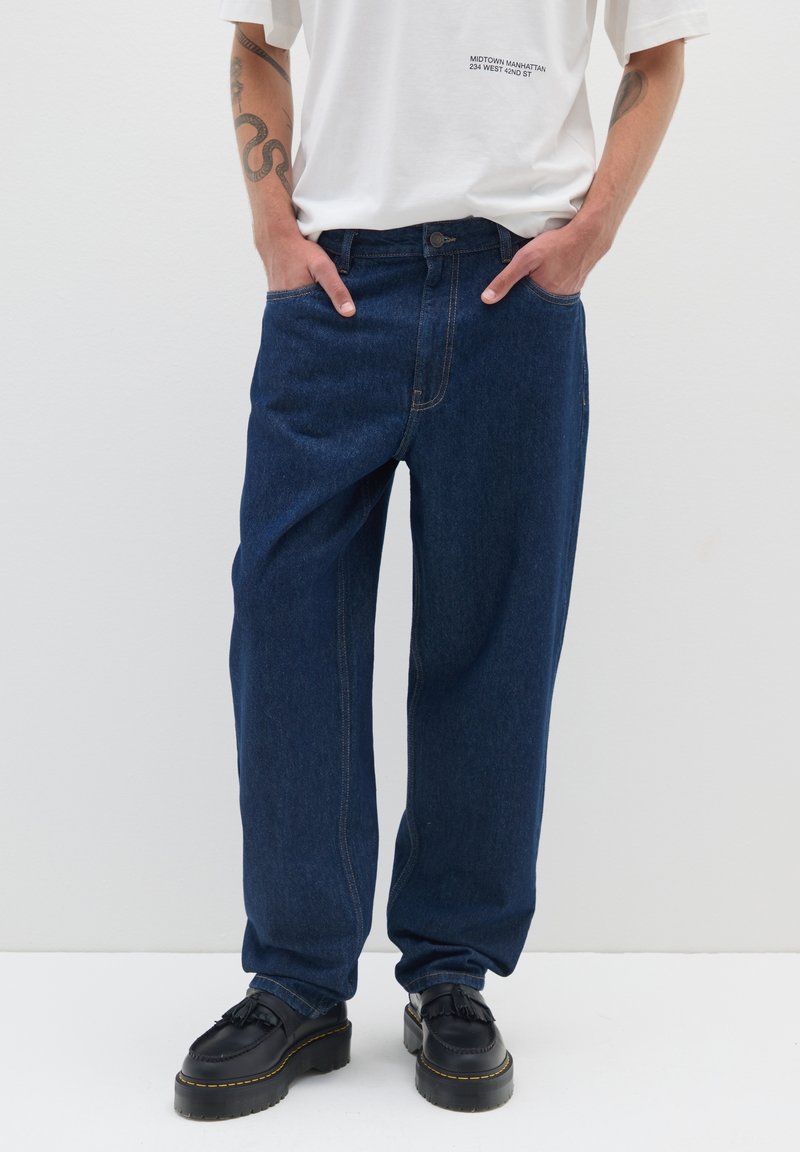 Jeans blu in denim con una vestibilità ampia, cuciture a contrasto e tasche laterali. Abbinati a scarpe nere. Maglietta bianca visibile sopra la vita.