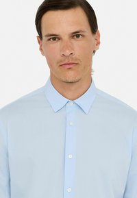 Camisa de botón azul claro con cuello estructurado y textura suave. Presenta una tapeta delantera con botones blancos y mangas largas.
