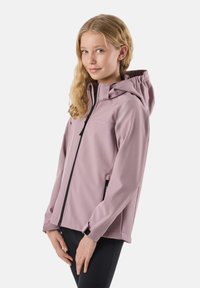 Mlada deklica, oblečena v vijoličasto jakno s kapuco in črnim zadrgo ter črne hlače, stoji s prepletenimi rokami spredaj in rahlo se smehlja.