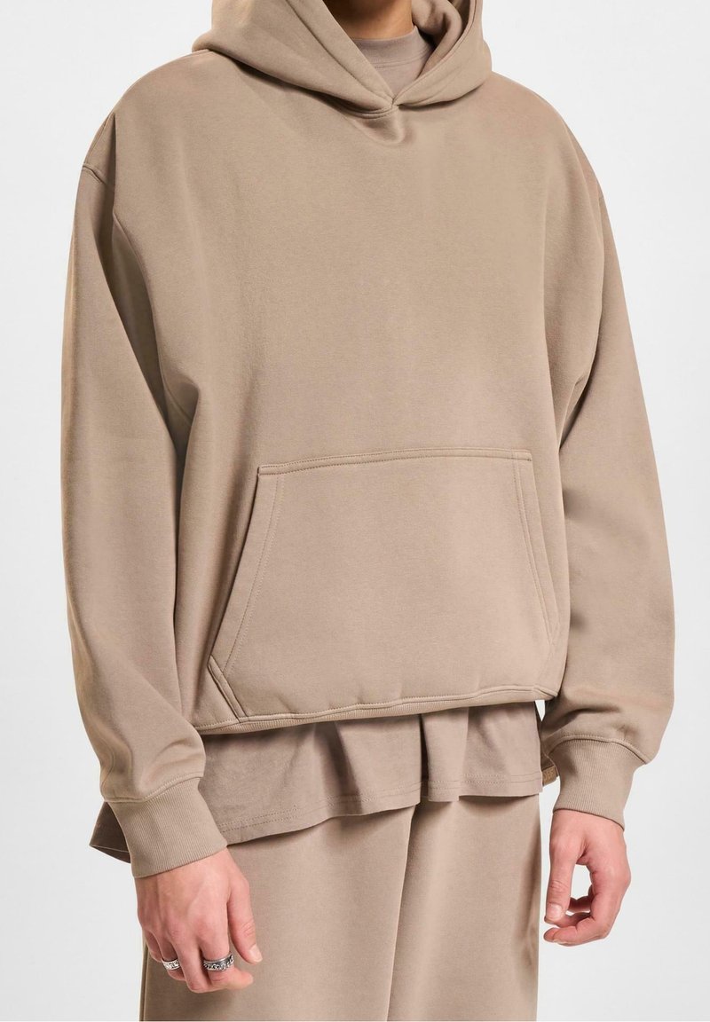 Homme portant un sweat à capuche beige surdimensionné avec poche avant, pantalon beige assorti et t-shirt beige superposé visible à l'ourlet.