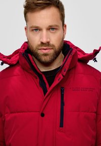 Rote gepolsterte Jacke mit hohem Kragen, Frontreißverschluss und Druckknopfverschlüssen. Verfügt über dunkle Akzente, eine Reißverschlusstasche und ein Branding auf der Brust.