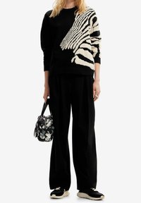 Kobieta w czarnym swetrze z dużym białym wzorem twarzy zebry, czarnych szerokich spodniach, czarnych trampkach i trzymająca małą torebkę w wzór.