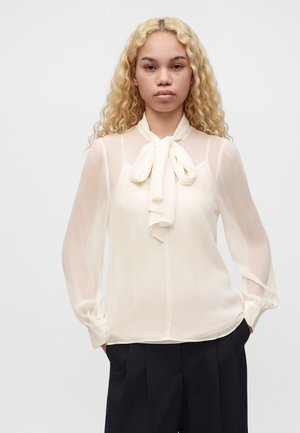 Jeune femme aux longs cheveux blonds bouclés porte un chemisier blanc transparent avec un col à nœud papillon et un pantalon noir taille haute, les mains dans les poches.