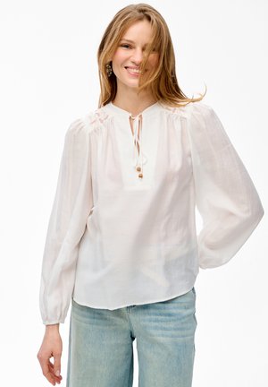 Kobieta o jasnobrązowych włosach, ubrana w białą bluzkę z długim rękawem z wiązaniem przy szyi oraz jasnoniebieskie dżinsy, uśmiechająca się na białym tle.