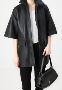 Abrigo corto de cuero negro con mangas cortas y cuello alto, con un cierre de botones en el frente y dos bolsillos delanteros. Acompañado de un bolso negro.