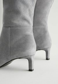Botas de tobillo de ante gris con un tacón cónico, que presentan una textura suave y un diseño sin costuras para una apariencia elegante. Detalles en negro en el tacón.
