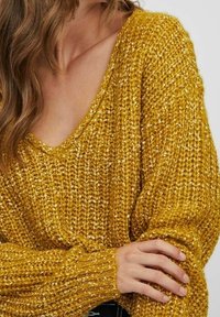 Gelbes Strickpullover mit V-Ausschnitt, lockerem Schnitt und gerippten Bündchen. Die Textur ist grob mit metallischen Fäden durchzogen.