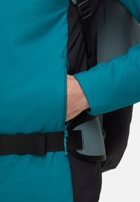 Veste teal avec des accents noirs, dotée d'une poche latérale, de détails zippés et d'une taille ajustable. Le matériau semble léger et durable.