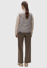 Vesta căptușită gri, cu cusături în formă de zigzag, purtată cu un top bej cu mânecă lungă și pantaloni maro, văzută din spate.