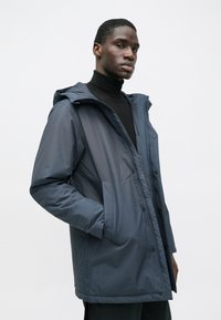 Ναυτικό μπουφάν με κουκούλα, φτιαγμένο από λείο υλικό, με φερμουάρ, κουμπιά σναπ και δύο πλάγιες τσέπες. Το σχέδιο είναι άνετης εφαρμογής.