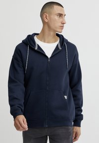 Marine blauwe hoodie met rits en verstelbare capuchon, zachte textuur, voorzakken en een klein label aan de zoom. Heeft contrasterende accenten met trekkoorden.