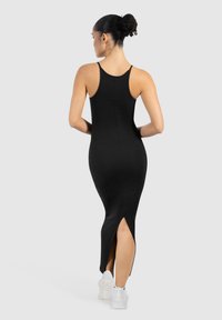 Kvinde med krøllet hår sat op i en knold iført en tætsiddende sort ærmeløs kjole med en slids bagpå, gående i hvide sneakers mod en ensfarvet baggrund.