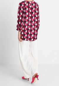 Blouse multicolore avec un motif géométrique, manches trois-quarts, fermeture boutonnée à l'arrière, associée à un pantalon large blanc et des talons roses.