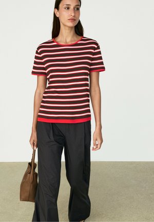 Frau, die ein rot-weiß-schwarz gestreiftes Kurzarmshirt, schwarze Hosen trägt, eine braune Handtasche hält und vor einem einfarbigen Hintergrund steht.