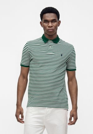 Homme portant un polo rayé horizontal vert et blanc avec un col vert et un pantalon blanc, debout devant un fond blanc uni.