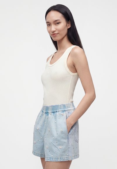 Femme aux longs cheveux foncés portant un haut sans manches blanc et un short en denim bleu clair avec broderies florales, debout les mains dans les poches.