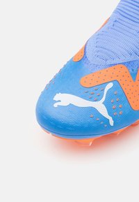 Blauwe voetbalschoenen met een oranje accentontwerp, voorzien van een gestructureerd oppervlak en een wit logo. Gemaakt van synthetisch materiaal voor grip.