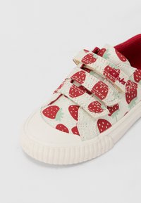 Canvas-Schuh mit einem weißen und roten Erdbeer-Muster, kontrastierendem roten Innenfutter und drei Klettverschlüssen. Weiße Gummisohle mit gerippter Textur.