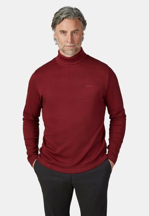 Middelbare man met grijs haar en baard draagt een rode coltrui en zwarte broek, staande met handen in de zakken tegen een witte achtergrond.